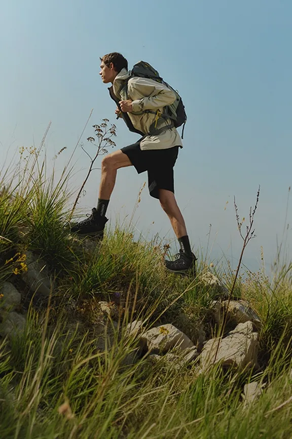 Ein Mann mit Rucksack wandert einen grasbewachsenen Hang unter blauem Himmel hinauf. 