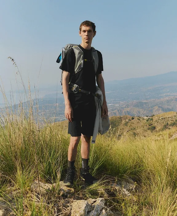 Ein Mann in Outdoor-Bekleidung steht mit Wanderrucksack auf einem Berg vor weitem Panorama.